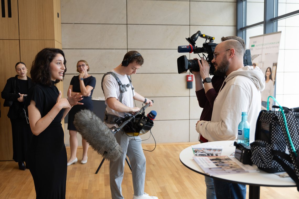 Eine Stipendiatin steht links im Bild und spricht zum Reporter-Team des WDR auf der rechten Seite.