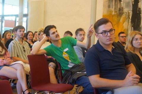 Das Foto zeigt Teilnehmerinnen und Teilnehmer der Veranstaltung, die in Stuhlreihen hinter- und nebeneinander sitzen und sich an einer Diskussion mit dem/der Vortragenden beteiligen, den/die man nicht auf dem Bild sieht.