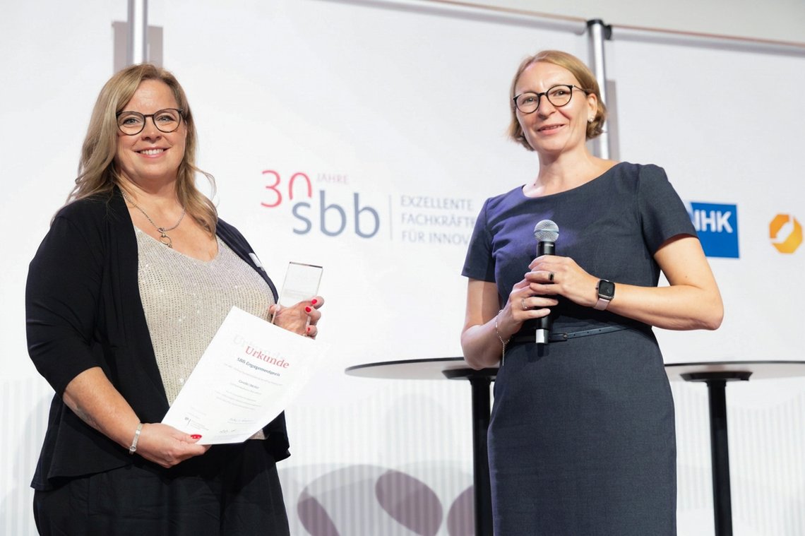 Carolin Weiler und Melanie Lochner stehen gemeinsam auf der Bühne. Frau Weiler steht links und hält den Preis und eine Urkunde in der Hand.