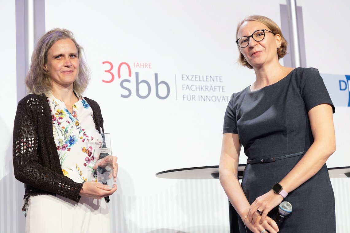Antje da Silva Santos und Melanie Lochner stehen gemeinsam auf der Bühne. Frau Weiler steht links und hält den Preis in der Hand.