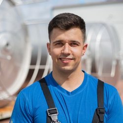 Portraitfoto des Stipendiaten Lukas Kriebel in Arbeitskleidung, im Hintergrund sieht man unscharfe große Behälter