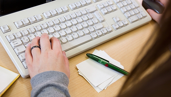 Foto: Hände auf Tastatur und Notizzettel mit Stift
